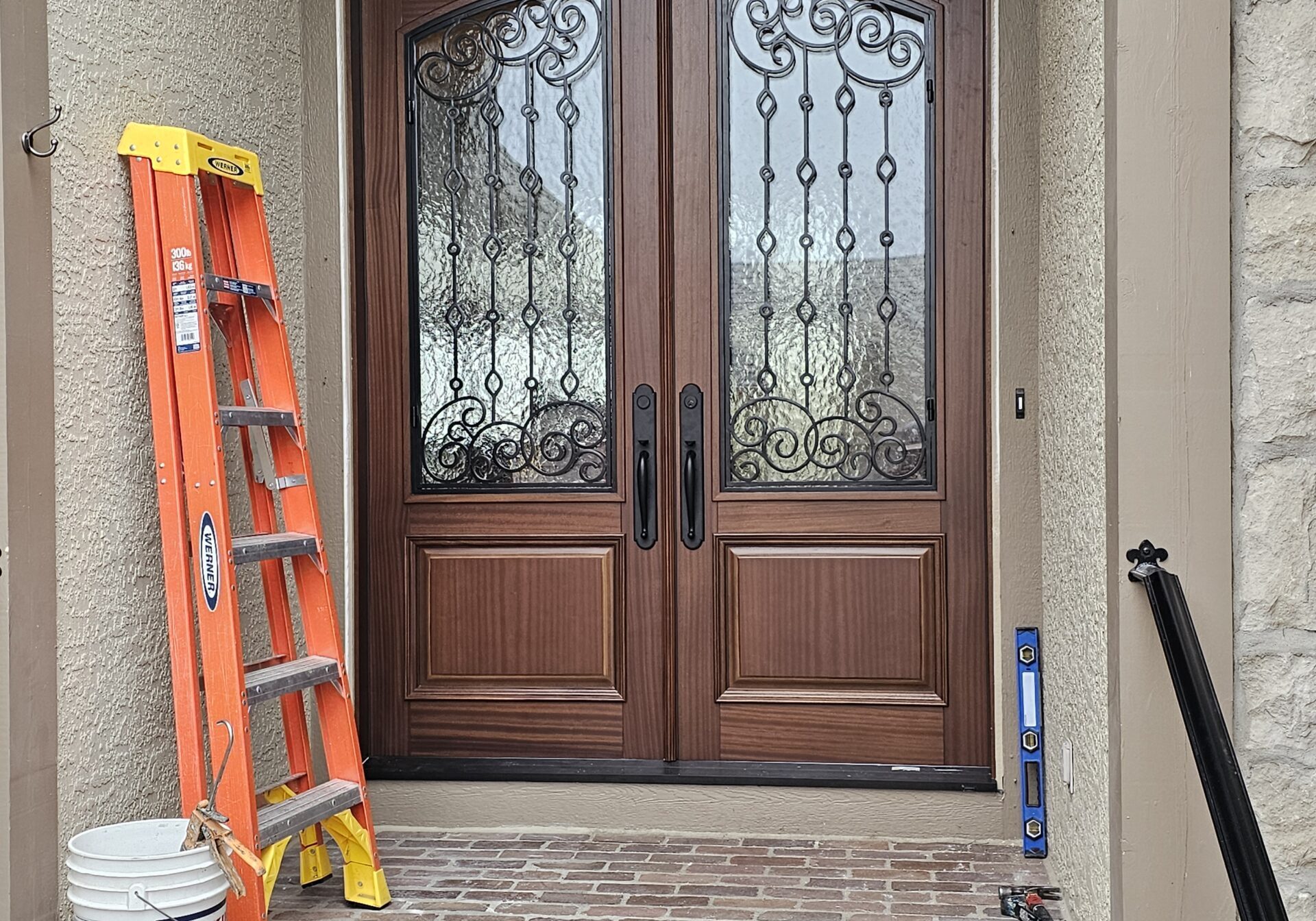 A decorative wood double front door with stone walls. On the left is an orange and yellow ladder leaning on the wall. Next to the ladder is a white bucket.