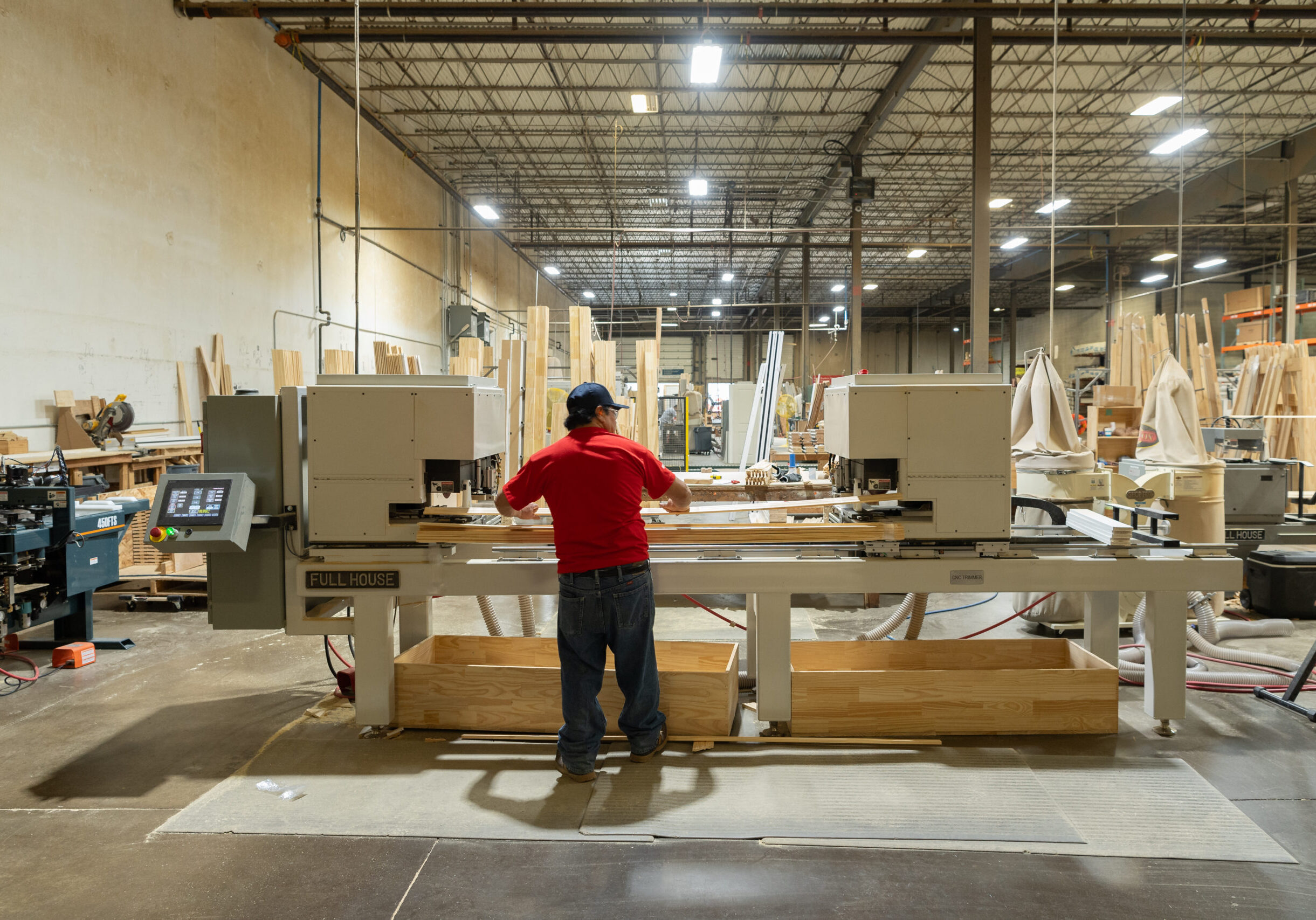 A man in a red shirt and blue pants wearing a baseball cap operates automatic milling machines in a large warehouse.